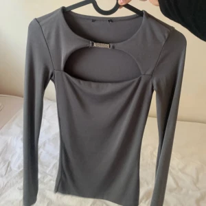 Grå långärmad topp med cut out - Säljer en stilren grå långärmad topp med snygg cut out-detalj vid bröstet och en liten glittrig detalj mitt fram. Toppen har en tight passform och är gjord i ett stretchigt material som sitter skönt på kroppen.