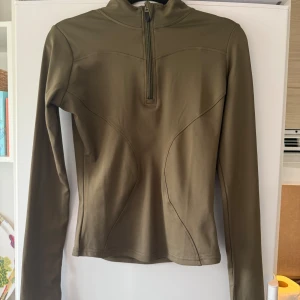 Olivgrön långärmad träningströja half zip - Olivgrön långärmad träningströja med half zip och hög krage. Snygga sömdetaljer framtill och tumhål vid ärmsluten. Aldrig använd