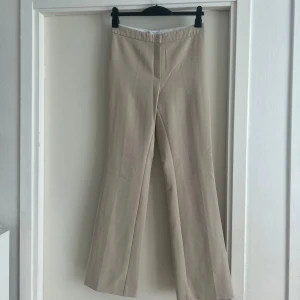 Beiga kostymbyxor med bootcut - Stilrena beiga kostymbyxor med bootcut-snitt och pressveck. Byxorna har hög midja och två dekorativa bakfickor. Perfekta för en clean och dressad look. Materialet är mjukt och känns bekvämt mot huden.