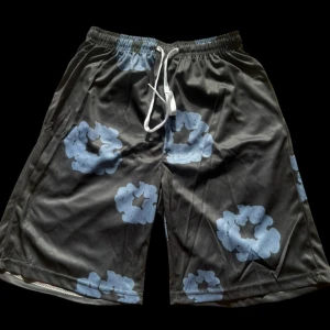 Svarta shorts med blått blommönster - Snygga svarta shorts med stora blå blommor och vit snörning i midjan. De har en loose passform och är perfekta för chill dagar. Materialet känns lätt och skönt mot huden, och midjan är elastisk för extra komfort.