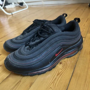 Nike Air Max 97 svart/röd sneakers - Nike Air Max 97 sneakers i svart med röda detaljer och reflekterande silverränder. Nypris 2400kr. Sulor saknas. Använd fåtal gånger.
