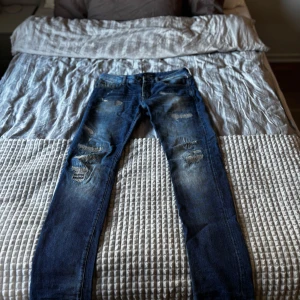 Blå slitna jeans från Scotch & Soda - Säljer ett par blå jeans från Scotch & Soda med coola slitningar och lagade detaljer på benen. Jeansen har klassisk femficksdesign, normal passform och är tillverkade i bomull. Perfekta för dig som gillar en avslappnad och trendig look. Skriv för fler frågor eller bilder.