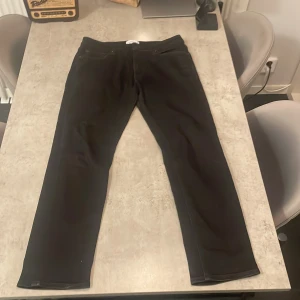 Svarta jeans Jack & Jones Regular Clark - Svarta jeans från Jack & Jones i modellen Regular Clark. Klassisk femficksdesign med raka ben och normal passform. Jeansen har svarta sömmar, bälteshällor och metallknappar. Materialet är jeans med en matt finish.