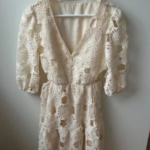 Beige spetsklänning med v-ringning - Superfin kort klänning i beige spets med v-ringning och puffiga trekvartsärmar. Klänningen har ett detaljerat virkat mönster och markerad midja. Perfekt för sommarens alla tillfällen såväl som student som midsommar! Liknar den populära ”majeklänningen”💓Har någon sminkfläck vid urringningen men inget som syns när man har den på sig💓