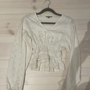 Vit blus med broderade blommor från Kiabi - Superfin vit blus från Kiabi i bomull med broderade blommor och v-ringning. Toppen har långa ärmar, smockad midja och söt knytning framtill. Perfekt för en fräsch och romantisk look med volangdetaljer och luftig känsla.