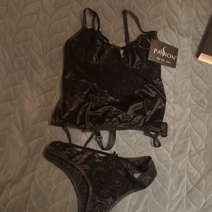 Svart korsettopp i satin från Passion - Snygg svart korsettopp i glansigt satinmaterial med spetsdetaljer och justerbara axelband. Toppen har dekorativa band och broderade blommor framtill. Perfekt för att skapa en cool och elegant look.