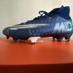 Nike SUPERFLY 7 ELITE MDS FG fotbollsskor - Snygga blå Nike SUPERFLY 7 ELITE MDS FG fotbollsskor med högt skaft och snörning. Skorna har mönstrad ovandel med vita och silverfärgade detaljer, samt robust dobbsula för gräsplan. De har inga sulor. De är lätta och smidiga med textil och syntetmaterial för bästa passform och grepp på planen. Perfekt för dig som vill vara så snabb som möjligt på planen.