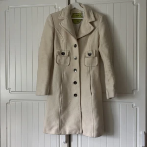 Beige kappa från Miss sixty med fickor - Snyggaste vita kappa från Miss sixty med markerad krage, två stora fickor framtill och svarta knappar med logga. Jackan är figursydd och har långa ärmar. Perfekt för dig som vill ha en stilren och trendig look under kallare månader. Ska vara storlek M men är liten i storleken så snarare en S