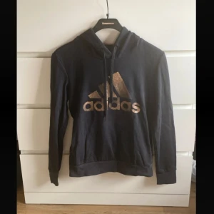 Svart hoodie från Adidas med guldtryck - Svart hoodie från Adidas i storlek XS med stort guldigt logotryck på bröstet. Tröjan har huva med snörning och långa ärmar. Perfekt för en sportig och chill stil. Mjuk insida och klassisk passform.