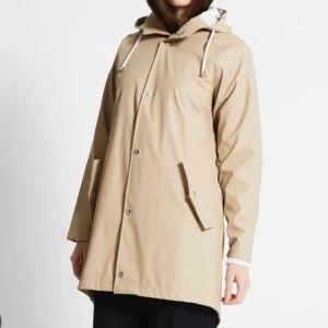 Beige regnjacka med huva - Stilren beige regnjacka med huva och vita dragsnören. Jackan har tryckknappar framtill, två snedställda fickor och en rak, något längre passform. Perfekt för regniga dagar när du vill hålla dig torr och snygg. (Säljs inte i denna färgen längre)