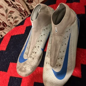 Nike fotbollsskor i vitt och blått - Säljer ett par Nike fotbollsskor i vitt med blå detaljer och stor Swoosh-logga på sidan. Skorna har en strömlinjeformad design utan synlig snörning och är tillverkade i syntetmaterial. Perfekta för dig som vill sticka ut på planen.