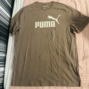 Brun t-shirt från Puma i bomull - Snygg brun t-shirt från Puma med klassisk logga i vitt framtill. T-shirten är i mjuk bomull och har rund hals och korta ärmar. Perfekt för en avslappnad och sportig stil.