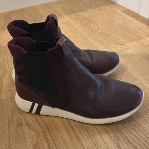 Vinröda chelsea boots från Gant - Snygga vinröda chelsea boots från Gant i skinn med elastiska svarta paneler på sidorna och vit platt sula med dekorativa ränder. Skorna har en sportig vibe och är perfekta för dig som vill ha något stilrent men ändå bekvämt.