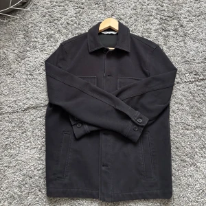Svart overshirt med knappar - Stilren svart overshirt med klassisk krage och knäppning framtill. Jackan har två bröstfickor och två sidofickor, samt långa ärmar med knappar vid ärmsluten. Perfekt för lager-på-lager och enkel att matcha.
