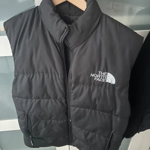  North Face - Säljer en svart dunväst från The North Face i storlek S. Västen har hög krage, dragkedja framtill och två fickor med dragkedja. Klassisk logga broderad på bröstet. Perfekt för lager-på-lager och riktigt snygg till streetwear den är fejk.