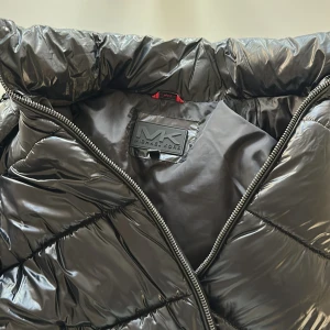 Svart pufferjacka från Michael Kors i nyskick - Snygg svart pufferjacka från Michael Kors med glansig finish och coola diagonala sömmar. Jackan har hög krage, dragkedja framtill och två fickor med dragkedja. Tillverkad i återvunnen polyester och designad för att vara både vind- och vattenavvisande. Jackan är använd ett fåtal gånger och är i nyskick 