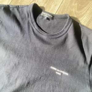Svart t-shirt Comme des Garçons Homme - Svart t-shirt från Comme des Garçons Homme med diskret vit logotyp på bröstet. Klassisk rund hals och korta ärmar. Tillverkad i 100% bomull, vilket gör den mjuk och skön att bära. Perfekt för dig som gillar minimalistisk och stilren design.