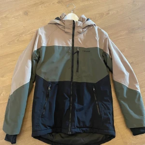 Skidjacka från Everest - Snygg skidjacka från Everest i färgblock med beige, olivgrön och svart. Jackan har huva, flera dragkedjefickor och justerbara ärmslut. Perfekt för kyliga dagar och har en sportig look med logga på bröstet. Den är i fint skick, hör av dig vid frågor😊
