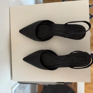 Open Edit, Svarta pumps med spetsig tå - Stilrena svarta pumps från Din Sko med spetsig tå och låg klack. Skorna har en öppen häl med smal rem runt ankeln och är tillverkade i syntetmaterial. Perfekta för dig som gillar en klassisk och elegant look.