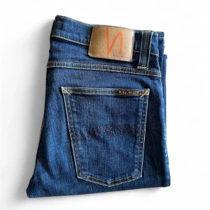 Blå jeans från Nudie Jeans Co - Snygga blå jeans från Nudie Jeans Co med klassisk femficksdesign och kontrastsömmar. Jeansen har normal passform och raka ben, tillverkade i slitstarkt denim med en djupblå tvätt. Perfekta för dig som gillar stilrena och tidlösa jeans - W27 L32 - Hör av er vid frågor eller funderingar
