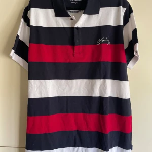 Salming pikétröja randig stl L - Snygg pikétröja från Salming i bomull med breda ränder i svart, vitt och rött. Klassisk krage med knappar och broderad logga på bröstet. Perfekt för en sportig och avslappnad stil.