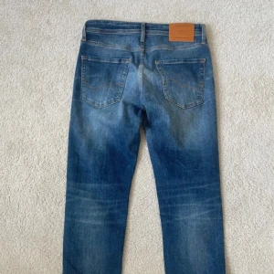 Blå jeans från Jack & Jones | W29 L30 - Snygga blå jeans från Jack & Jones med klassisk femficksdesign och ljusa slitningar framtill. Modellen heter Comfort/Mike och har raka ben med normal passform. Jeansen har bruna sömmar och läderpatch bak i midjan. Perfekta för en avslappnad stil.