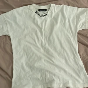 Vit Palm Angels t-shirt med tryck - Vit t-shirt från Palm Angels i bomull med svart logotyp-tryck både på kragen och stort över ryggen. Klassisk passform och korta ärmar, perfekt för en clean streetwear-look.