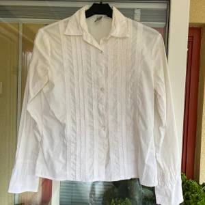 Vit skjorta från City DKNY med detaljer - Klassisk vit skjorta från DKNY med snygga vertikala broderade detaljer framtill. Skjortan har krage, långa ärmar med dubbla knappar vid manschetten och är tillverkad i bomull. 