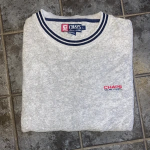 Grå långärmad tröja Chaps Ralph Lauren - Grå Chaps Ralph Lauren sweatshirt Riktigt snygg vintage-tröja med broderad logga på bröstet. Har en klassisk rund hals med blå/vita ränder och matchande muddar som gör den lite extra speciell.  Supermjuk och bekväm – perfekt till jeans eller baggy cargos. Funkar både som vardagsplagg och om du vill ha den där laid back streetwear-looken.