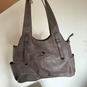 Väska - Stilren väska i vintage stil. Väskan har flera fack, både invändigt och utvändigt, samt justerbar axelrem och snygga metalldetaljer. Perfekt storlek för att få plats med allt du behöver. Kommer inte till användning hos mig tyvärr.