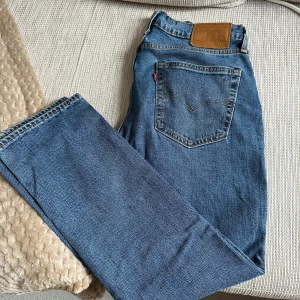 Levi's 514 blå jeans straight fit - Klassiska blå Levi's 501 jeans med rak passform och femficksdesign. Jeansen har den ikoniska läderpatchen bak i midjan, synliga nitar och dragkedja. Tillverkade i slitstarkt denim med snygg tvätt och orange sömmar. Har en lagning vid skrevet, se andra bilden.