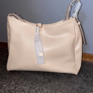 Beige axelväska från Anna Field - Nyskick/Helt oanvänd. Produkten är slutsåld på hemsidan. Originalpris 399kr. Stilren beige axelväska från Anna Field i mjukt syntetmaterial. Väskan har en enkel, slät design med en dekorativ knutdetalj framtill och bred axelrem. Perfekt storlek för att få plats med allt du behöver till vardagen.Utvändigt material: Polyuretan Foder: Textil Tyg: Skinnimitation  Höjd: 23 cm i storlek One Size Längd: 33 cm i storlek One Size Vidd: 12 cm i storlek.  Fack: Mobiltelefonfack Förslutning: Magnetlås