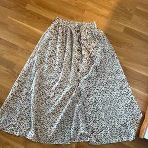 Leopardmönstrad långkjol med knappar - Snygg långkjol med leopardmönster i svart och beige. Kjolen har resår i midjan och knappar framtill för en cool detalj. Perfekt för dig som vill sticka ut lite extra med ett djurmönster.