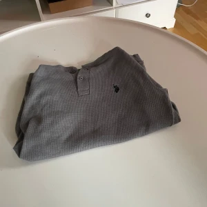 Ralph lauren tröja long sleeve  - Grå långärmad tröja från U.S. Polo Assn. med knappslå vid halsen och broderad logga på bröstet. Tröjan är i ett mjukt, ribbat material av bomull och polyester, perfekt för chill dagar. Enkel och stilren design som funkar till det mesta.