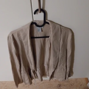 Beige kavaj från H&M, strl 40 - Snygg beige kavaj från H&M i storlek 40. Jackan har en avslappnad passform med knytband i midjan och långa ärmar. Perfekt för dig som gillar en stilren och enkel look. Materialet känns mjukt och lätt, vilket gör den skön att bära.