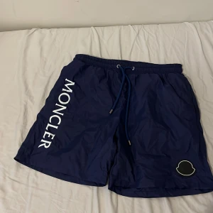 Marinblå Moncler shorts med snörning - Snygga marinblå shorts från Moncler med vit logotyp längs benet och en rund Moncler-patch. De har elastisk midja med snörning och metalländar med Moncler-gravyr. Materialet är lätt och syntetiskt, perfekt för sommardagar eller bad.