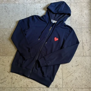 Mörkblå Cdg play zip - Snygg mörkblå hoodie från Cdg Play med dragkedja och huva. Tröjan har den ikoniska röda loggan broderat på bröstet. Storlek S och är i toppskick. Fraktas inom 24h✅