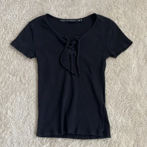 Svart ribbad topp med snörning - Svart ribbad topp från lager 157 i storlek M. Toppen har korta ärmar och snygg snörning framtill vid halsringningen. Materialet är stretchigt och sitter tight.