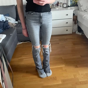 Grå slitna jeans från Zara, strl 36 - Midjemått: 31,5cm Innerbensmått: 81cm Ytterbensmått: 110cm        Grå jeans från Zara i storlek 36 med snygga slitningar och hål över knäna. Modellen har raka ben och klassisk femficksdesign. Jeansen har hög midja och är tillverkade i bomull med en cool, tvättad look.