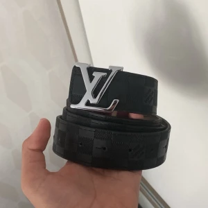 Svart Louis Vuitton bälte med rutigt mönster - Snyggt svart bälte från Louis Vuitton med klassiskt rutigt mönster och en stor, silverfärgad LV-logga som spänne. Bältet är gjort i skinn och har en stilren, modern look som passar till många outfits.