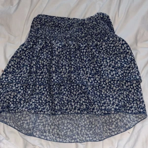 Blå blommig kortkjol med resår - Supersöt blå kortkjol med vitt blommönster och resår i midjan. Kjolen har en luftig och flowy känsla, perfekt för varma dagar. Den är lätt och har en volangig nederdel som ger extra rörelse. Passar perfekt till sneakers eller sandaler.