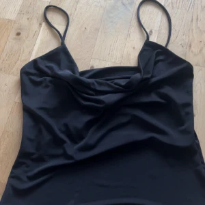 Svart satinlik fest topp från H&M - Svart festtopp från H&M med tunna axelband och snygg draperad urringning. Bak så är den nästan helt öpppen förutom bandet som håller ihop den. Den är lite urringad i fram. 