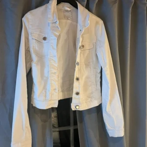 Vit jeansjacka med silvriga knappar - Snygg vit jeansjacka med klassisk krage, två bröstfickor och silvriga knappar framtill. Jackan är croppad och har långa ärmar. Perfekt för dig som vill ha en fräsch och stilren look till våren eller sommaren. Är en fläck på fickan går säkert bort med vanish har inte testat