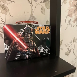 Star Wars plåtburk med Darth Vader - Cool plåtväska med Star Wars-tryck, Darth Vader och Stormtrooper på sidorna. Väskan är svart, röd och silver med rött handtag och detaljer i metall. Perfekt för att förvara småprylar eller som lunchlåda. Motiv och logga på både framsida och baksida.