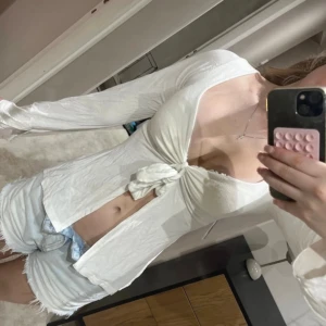 Vit långärmad omlottopp med knyt - Säljer en vit långärmad topp med snygg omlottdesign och knyt framtill. Toppen har djup urringning och är croppad, vilket ger en trendig och fräsch look. Perfekt att matcha med jeans eller shorts för en cool stil.