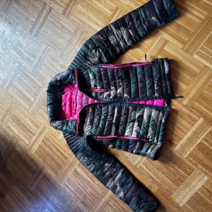 Superdry camo pufferjacka med rosa detaljer - Säljer en pufferjacka från Superdry med camouflage-mönster i grönt, brunt och beige. Insidan är knallrosa och jackan har rosa dragkedjor. Jackan är quiltad och har lång ärm, perfekt för dig som vill sticka ut med coola färgkontraster.