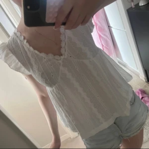 Vit spetsblus med volangärmar - Supersöt vit blus med spetsdetaljer och v-ringning. Blusen har volangärmar och ett broderat mönster med små hål och geometriska former. Perfekt för varma dagar och ger en romantisk vibe till din outfit.