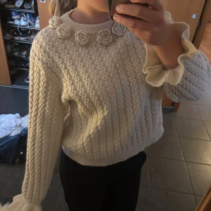 Beige stickad tröja med volangärm - Superfin beige stickad tröja med vågigt mönster och dekorativa virkade blommor runt halsen. Tröjan har långa ärmar med volangdetalj vid ärmslut och ribbade muddar. Perfekt för dig som gillar unika detaljer och mysig stil.