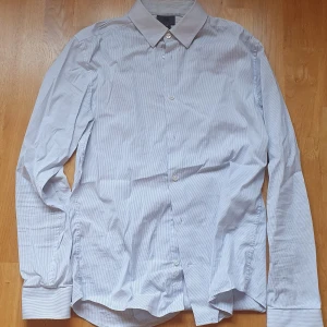 Vit och blå-randig skjorta från H&M - Säljer en slim fit skjorta från H&M i vitt med tunna blå ränder. Skjortan har klassisk krage, långa ärmar med manschett och knäppning framtill. Materialet är mjukt och stretchigt med 75% bomull, 22% polyamid och 3% elastan. Väldigt lite använd.