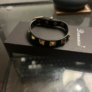 Liknade valentino armband  - Coolt svart armband från Demasi i skinn med guldfärgade pyramidnitar runt om. Spännet är också i guldton och armbandet har justerbar längd. Perfekt accessoar för att ge din outfit en edgy vibe.